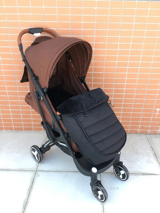 Une poussette marron avec un couvre-pieds noir WarmCott™ Chancelière pour bébé est posée sur un sol carrelé devant un mur en briques couleur pêche. Elle est équipée de quatre roues et d'un élégant guidon marron.