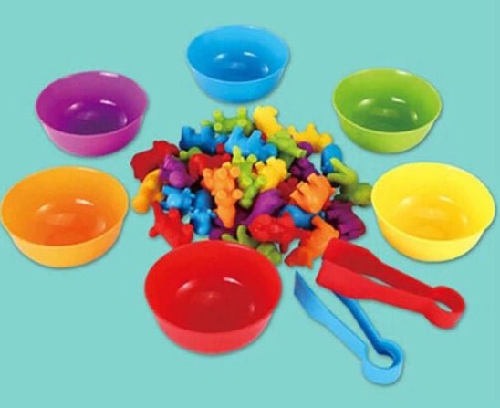 Six bols en plastique colorés entourent une pile de Figurine animaux - Jouet bebe, chaque bol étant d'une couleur différente. Une pince à épiler en plastique bleu se trouve à proximité, ce qui fait ressortir ce jouet éducatif sur le fond sarcelle.