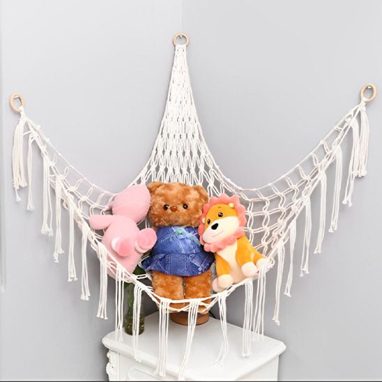 Le Hamac peluche - Jouet bebe est accroché dans le coin d'une pièce gris clair, avec un ours rose, un ours brun en salopette bleue et un lion jaune au-dessus d'une petite table blanche où sont rangés d'adorables jouets en peluche.