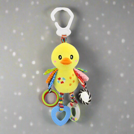 La Peluche - Jouet pour bébé est un oiseau en peluche jaune avec un bec orange, des ailes multicolores et des anneaux et formes en plastique suspendus. Il se fixe à l'aide d'un clip en plastique blanc et présente un fond gris avec des points blancs pour la stimulation sensorielle.