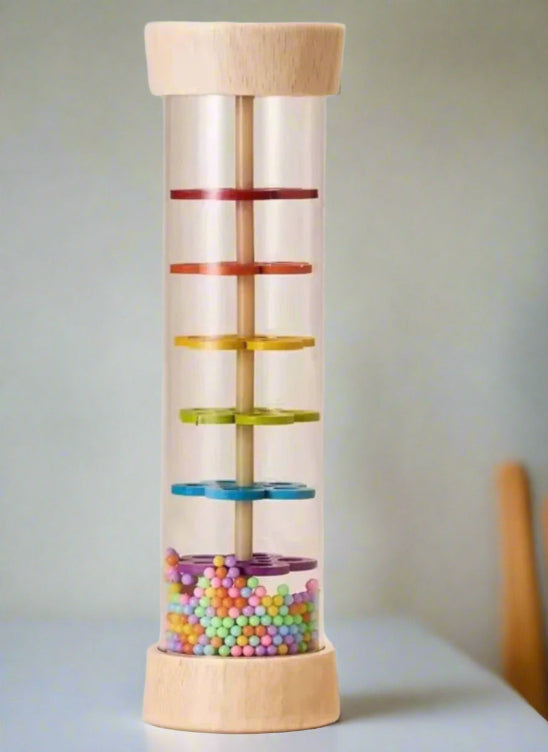 Rain Sound -Jouets en bois est un bâton de pluie cylindrique transparent avec un couvercle et une base en bois, rempli de perles colorées et de six disques multicolores empilés sur une tige centrale. Les perles s'accumulent au fond du cylindre.