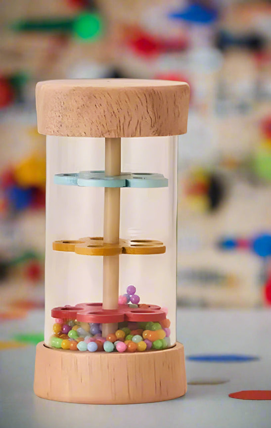 Rain Sound - Jouets en bois est un hochet cylindrique en bois avec des côtés transparents, trois disques colorés et des perles multicolores à l'intérieur. Il se tient debout sur une surface plane, sur un fond flou.