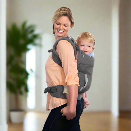 À l'intérieur, une femme souriante porte un jeune enfant sur son dos dans le Porte Bébé | Unisexe. L'arrière-plan légèrement flouté comprend une plante et des murs clairs.