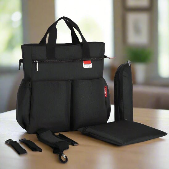 Le Sac a Langer - Longchamps & Poussette, un sac à langer noir avec de multiples poches et une étiquette rouge et blanche, est présenté sur une table avec une pochette assortie, un coussin rectangulaire et deux bandouliÚres détachables dans un décor intérieur légÚrement flou.