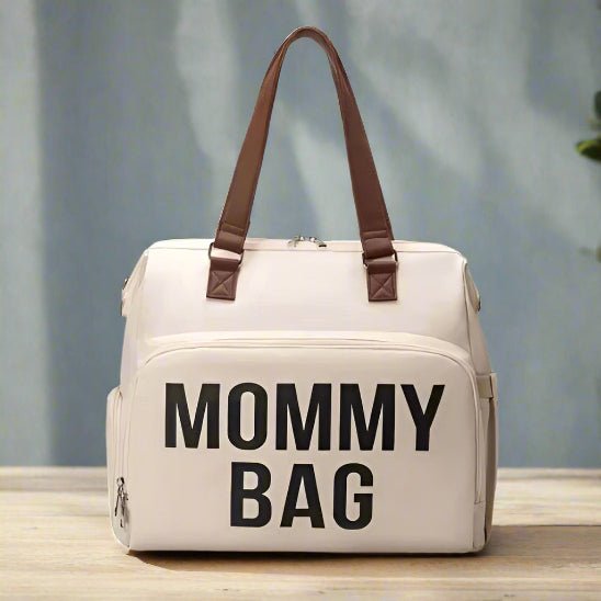 Le Sac a Langer | Mommy Bag, de couleur crÚme avec des poignées brunes et portant la mention "MOMMY BAG" en caractÚres noirs gras sur le devant, est présenté sur une surface en bois sur un fond bleu clair avec une partie d'une plante verte visible.