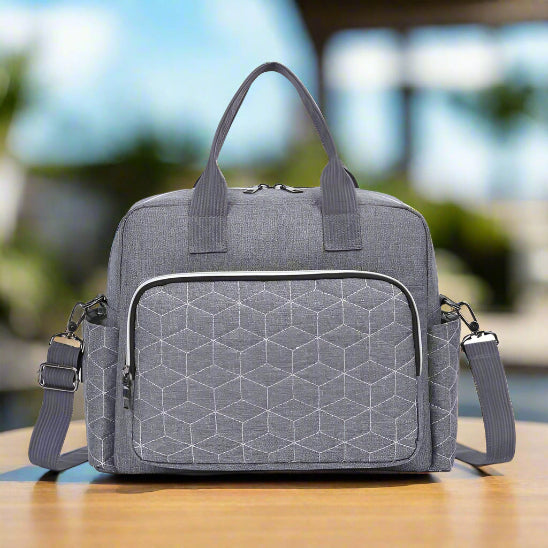 A Sac bebe | Sac de Maternité gris sac à langer avec une poche géométrique sur le devant repose sur du bois. Il présente une grande capacité, deux anses courtes, une bandouliÚre amovible et un compartiment principal zippé. Le fond est flou.