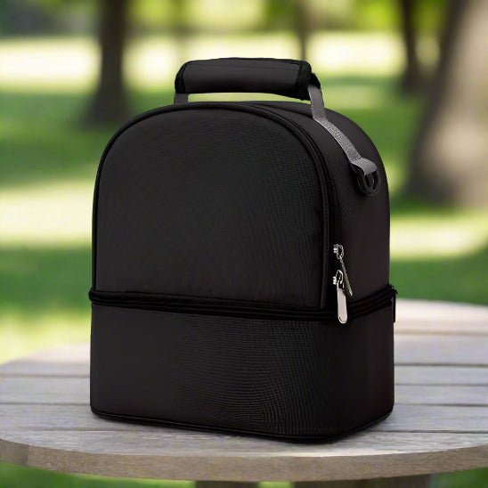 Le Petit Sac Bebe - Isotherme, un sac thermique noir élégant avec poignées et fermeture éclair, est posé sur une table ronde en bois à l'extérieur - idéal pour transporter des bouteilles ou des en-cas - avec un parc vert flou en arriÚre-plan.