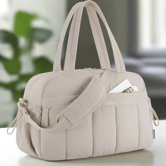 Le Sac a Langer - Longchamps, un sac de sport matelassé beige clair avec deux poignées, une bandoulière et des poches extérieures, dont l'une contient une enveloppe blanche, est présenté sur une surface blanche à l'intérieur.