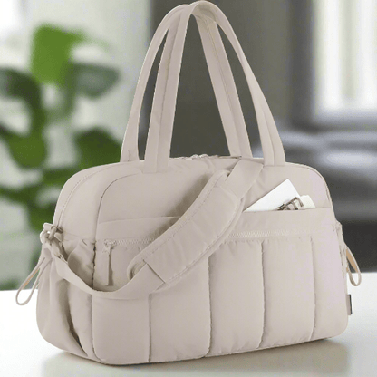 Le Sac a Langer - Longchamps, un sac de sport matelassé beige clair avec deux poignées, une bandoulière et des poches extérieures, dont l'une contient une enveloppe blanche, est présenté sur une surface blanche à l'intérieur.
