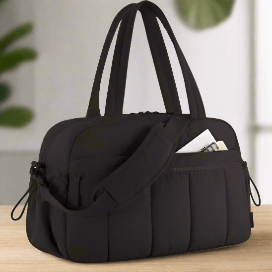 Le Sac à Langer de Longchamps, un sac à langer multifonctionnel matelassé noir avec double poignée et bandoulière, est posé sur du bois. Une bouteille d'eau blanche se trouve dans sa poche latérale, avec des plantes vertes floues et un fond blanc à l'arrière-plan.