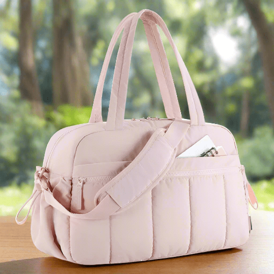 Le Sac à Langer - Longchamps en rose clair, avec un matelassage, deux anses et une bandoulière, repose sur une surface en bois. Un carnet et un stylo sont visibles dans la poche extérieure, sur fond vert flou.