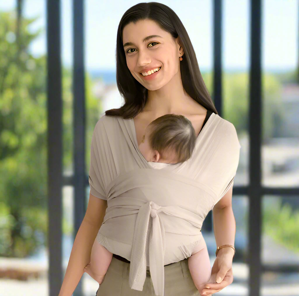 Une femme sourit à l'objectif tout en tenant un bébé dans le Porte-Écharpe Ergonomique, démontrant ainsi le portage ergonomique. De grandes fenêtres avec une vue floue sur l'extérieur sont visibles en arrière-plan.