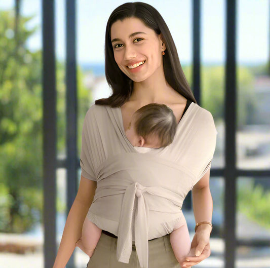 Une femme sourit à l'intérieur, portant son bébé dans le Porte-Écharpe Ergonomique sur sa poitrine. La lumière du soleil provenant des fenêtres floues met en valeur le confort naturel et le soutien ergonomique de cette écharpe de portage.