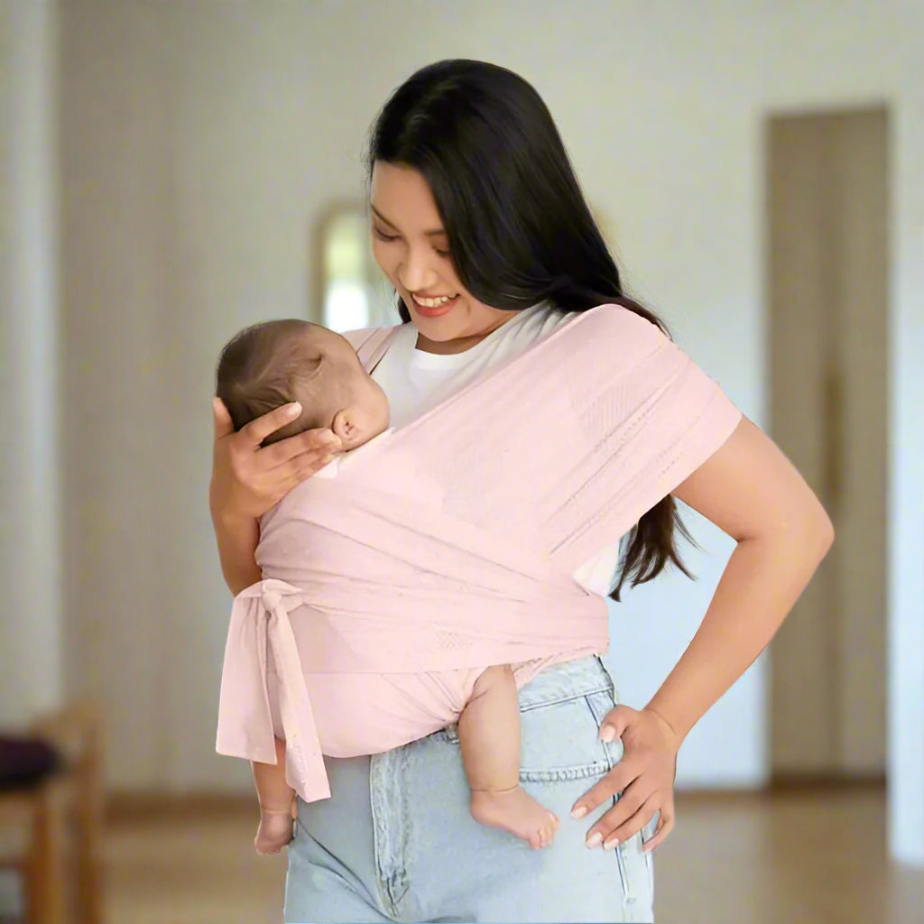 Une femme sourit à l'intérieur, tenant son bébé contre elle grâce au Porte-Écharpe Ergonomique, une écharpe de portage rose clair. Le bébé est confortablement installé face au corps, tandis que la femme le soutient d'une main.