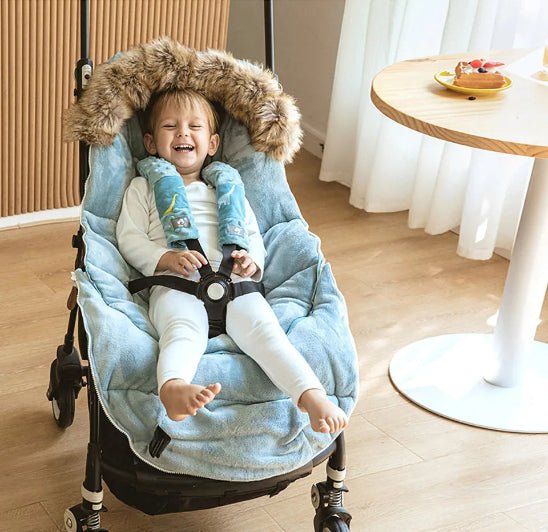 Un tout-petit profite du confort de la Chancelière | 0-36 mois, assis dans une poussette avec une couverture bleue et une capuche bordée de fourrure près d'une table ronde en bois avec de la nourriture et un verre, restant bien au chaud et protégé à l'intérieur.