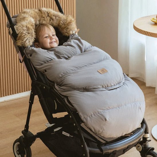 Un jeune enfant est installé dans une poussette, confortablement installé dans la Chancelière | 0-36 mois pour une protection thermique optimale. La poussette est posée sur un plancher en bois, près d'une table ronde et d'une fenêtre ornée de rideaux blancs transparents.