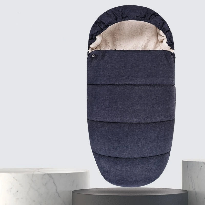 La Chancelière Bébé - Care+, une chancelière pour bébé rembourrée gris foncé avec une doublure en polaire de laine d'agneau de couleur crème, est présentée debout sur un fond clair au-dessus de marbre arrondi et de piédestaux sombres pour une protection thermique optimale.