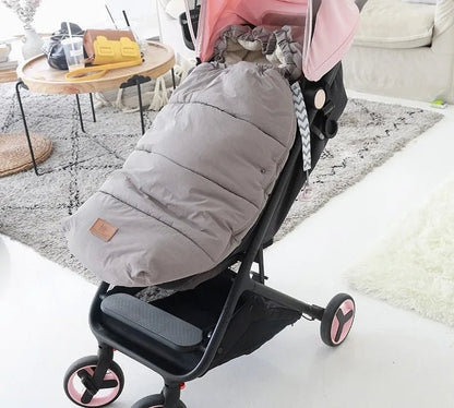 Une poussette aux accents roses et noirs est garée à l'intérieur sur un tapis clair, ornée de la Chancelière Bébé - Care+ grise et rembourrée pour une protection thermique. Une table ronde en bois avec divers objets apparaît en arrière-plan.