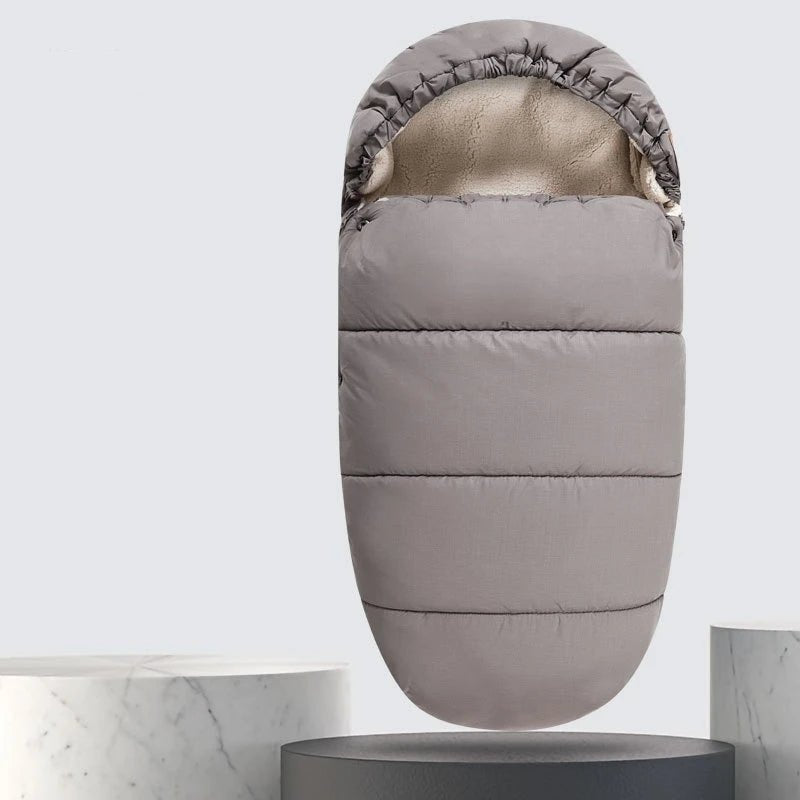 La Chancelière Bébé - Care+ est une chancelière pour bébé grise à capuche avec une doublure polaire pour une excellente protection thermique, présentée debout sur un piédestal rond à côté d'une surface texturée en marbre.