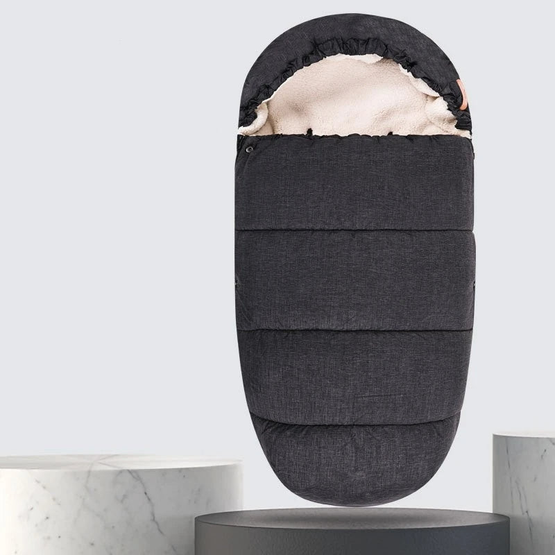 La Chancelière Bébé - Care+ en gris foncé, dotée d'une douce doublure en laine d'agneau crème, est présentée debout sur un socle rond avec des fonds clairs et des surfaces rondes, soulignant sa protection thermique.