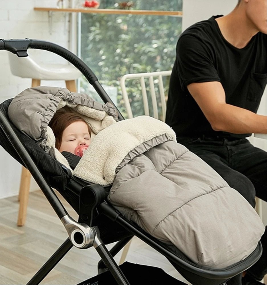 Un bébé dort dans une poussette, emmitouflé dans la Chancelière Bébé - Care+ pour une protection thermique optimale, avec une tétine. Un adulte en chemise noire est assis à côté, à une table, dans une pièce lumineuse aux grandes fenêtres.