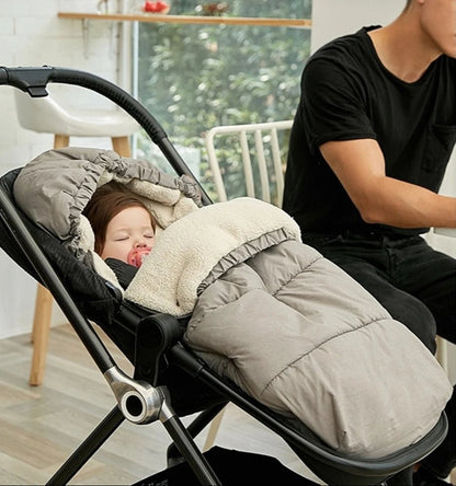 Un bébé dort dans une poussette, emmitouflé dans la Chancelière Bébé - Care+ pour une protection thermique optimale, avec une tétine. Un adulte en chemise noire est assis à côté, à une table, dans une pièce lumineuse aux grandes fenêtres.