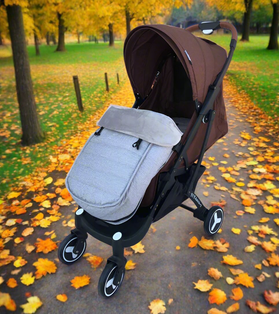 Une poussette marron et grise est garée sur un chemin de parc avec de l'herbe, des arbres et des feuilles d'automne, dotée de la chancelière bébé WarmCott™ pour une protection hivernale supplémentaire.