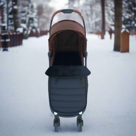 Une poussette avec une capote marron et une chancelière bébé WarmCott™ noire est garée sur un chemin enneigé bordé d'arbres et de clôtures, offrant une excellente protection hivernale en cette froide journée d'hiver.