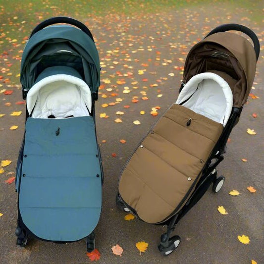 Deux poussettes sont assises côte à côte sur un chemin parsemé de feuilles. La gauche est vert foncé avec une chancelière bébé bleue, tandis que la droite est marron avec une Chancelière Bébé | Imperméable marron assortie. Les deux sont dotés d'un rembourrage blanc doux à l'intérieur.