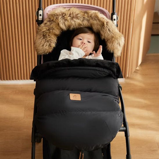 Un bébé dans une poussette, bien au chaud et protégé dans une Chancelière Poussette | 0-24 mois avec capuche bordée de fourrure, porte un haut léger et est assis près de panneaux muraux en bois et d'un sol clair.