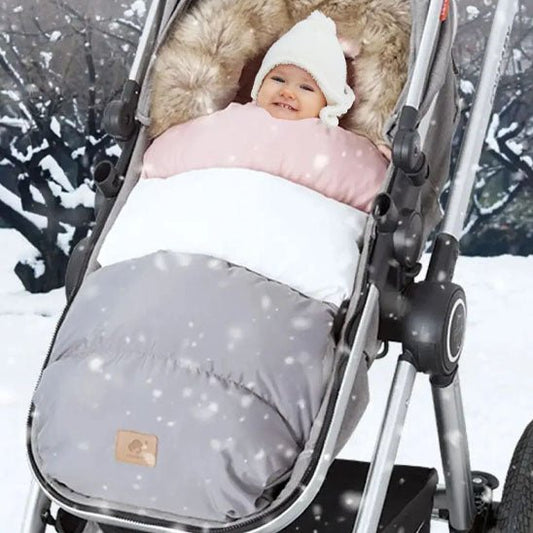 Un bébé dans un bonnet en tricot blanc est bien au chaud dans la Chancelière Poussette | 0-36 mois (rose, blanc, gris) pour une isolation thermique optimale, assis à l'extérieur dans un paysage enneigé avec des arbres tandis que de doux flocons de neige tombent pour le confort de bébé.