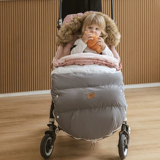 Un jeune enfant est assis dans une poussette, confortablement installé dans la Chancelière Poussette | 0-36 mois avec une excellente isolation thermique, blotti sous une couverture moelleuse grise et blanche avec une capuche bordée de fourrure, savourant une collation sur un plancher en bois avec un mur en bois nervuré derrière.