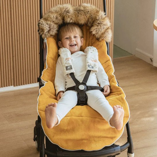 Un jeune enfant en blanc est assis en souriant dans une poussette avec la Chancelière Poussette | 0-36 mois, dotée d'une capuche en fourrure et de sangles de sécurité pour le confort et l'isolation thermique, à l'intérieur sur un plancher en bois près d'un mur nervuré.