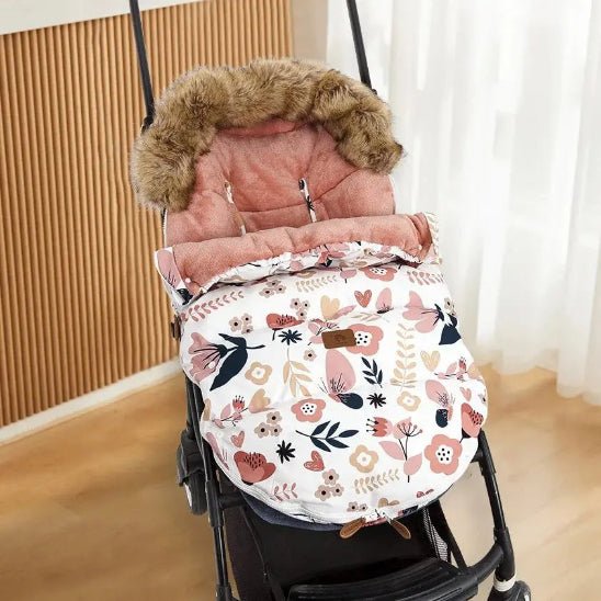 La Chancelière Poussette | Hiver 0-24 mois en rose et blanc fleuri, avec doublure capuche en fausse fourrure pour une protection thermique optimale, est placée à l'intérieur près d'un mur en bois et d'un rideau blanc transparent.