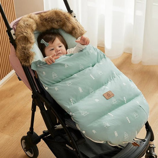 Un bébé est allongé dans une poussette, emmitouflé dans la Chancelière Poussette | Hiver 0-24 mois, vert clair à motifs et dotée d'une capuche en fausse fourrure pour un confort optimal. La poussette est posée à l'intérieur, sur un parquet, près de rideaux blancs transparents.