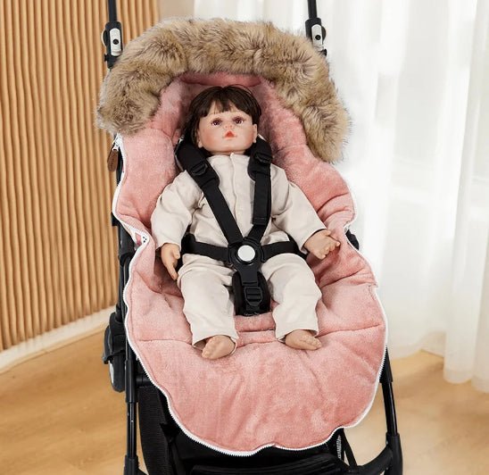 Une poupée bébé réaliste dans une tenue légère est fixée avec des sangles noires dans une poussette doublée de la Chancelière Poussette | Hiver 0-24 mois, une couverture bordée de fourrure rose qui offre une protection thermique et un confort supplémentaire pour bébé grâce au rideau et à la paroi en bois.