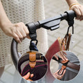 Une personne saisit la poignée d'une poussette avec deux crochets en cuir essentiels. L'image principale montre la poignée, tandis que les encarts mettent en évidence les détails : l'un comprend une sangle en cuir marron et un clip en métal noir, et un autre montre qu'il est ajusté sur un Crochet Pour Poussette réglable.