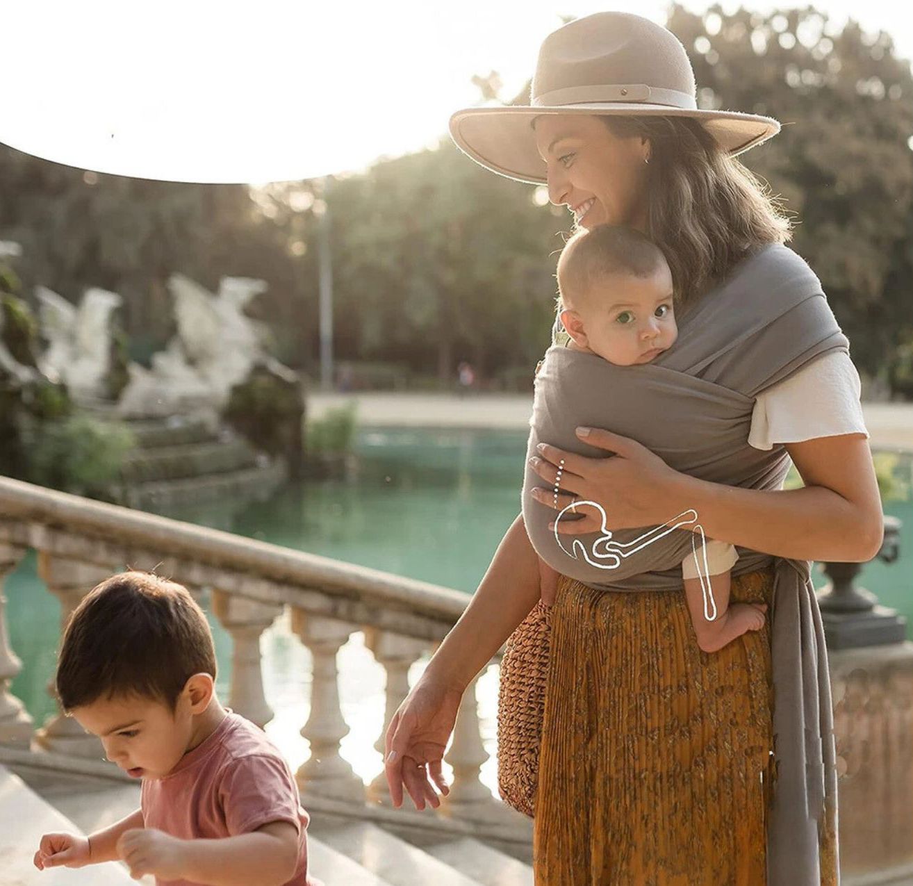 Une femme portant un chapeau se tient debout, près d'un étang, dans une écharpe de portage, portant son bébé de 0 à 36 mois, tout en tenant par la main un jeune enfant qui marche à ses côtés. Le bébé est tourné vers l'avant ; les deux enfants ont les cheveux bruns courts.