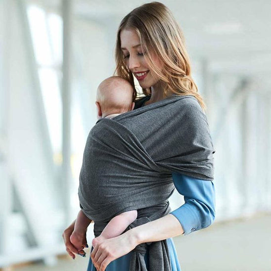 Une femme en robe bleue serre contre elle un bébé dans une écharpe de portage en coton gris ElasticFit™. Le bébé est tourné vers l'intérieur, dans un intérieur lumineux et moderne.