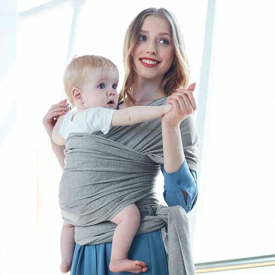 Une femme en robe bleue porte un bébé dans l'écharpe de portage bébé | ElasticFit™, en coton naturel. Le bébé, tourné vers l'extérieur grâce au soutien ElasticFit™, semble satisfait, tandis qu'ils regardent tous deux droit devant eux sous une lumière naturelle éclatante.