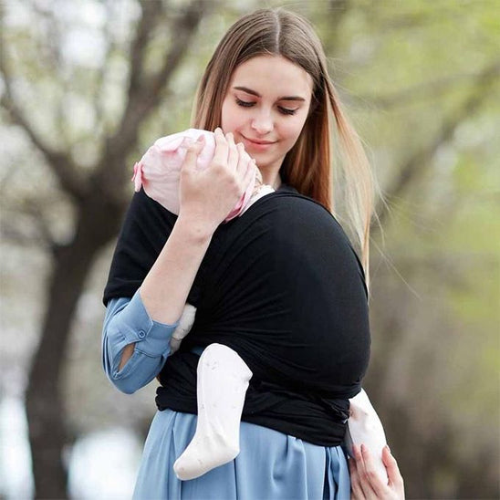 Une femme en robe bleue tient son bébé à l'extérieur, bien emmitouflé dans l'Écharpe de Portage Bébé | ElasticFit™ en coton naturel. Le bébé, vêtu de blanc, repose confortablement, tandis que des arbres verts ornent l'arrière-plan.