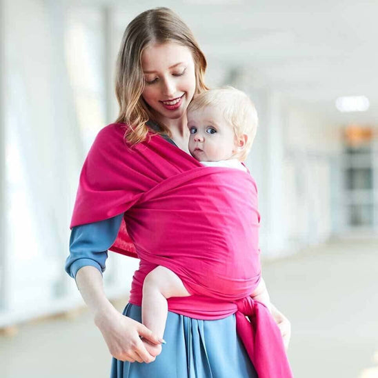 Une femme souriante, vêtue d'une robe bleue, porte son bébé face vers l'avant dans l'Écharpe de Portage Bébé | ElasticFit™, en coton naturel rose vif. L'arrière-plan légèrement flou suggère qu'ils sont à l'intérieur.