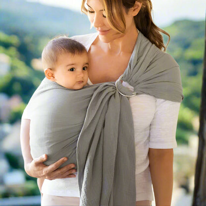 Une femme en haut blanc porte son bébé dans le porte-bébé gris Écharpe portage bébé | RingSling™. Le bébé apprécie le contact peau à peau, regarde vers l'avant et regarde de côté, dans un décor de verdure luxuriante aux douces nuances floues.