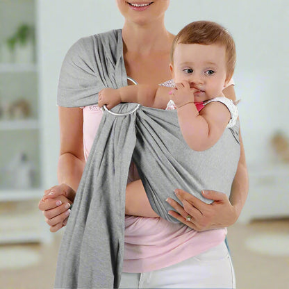 Une femme vêtue d'une chemise rose clair porte un bébé dans une écharpe de portage bébé grise | RingSling™ sur sa poitrine. Le bébé apprécie le contact peau à peau, suçant son pouce, tandis qu'ils se détendent à l'intérieur, avec des étagères floues derrière eux.