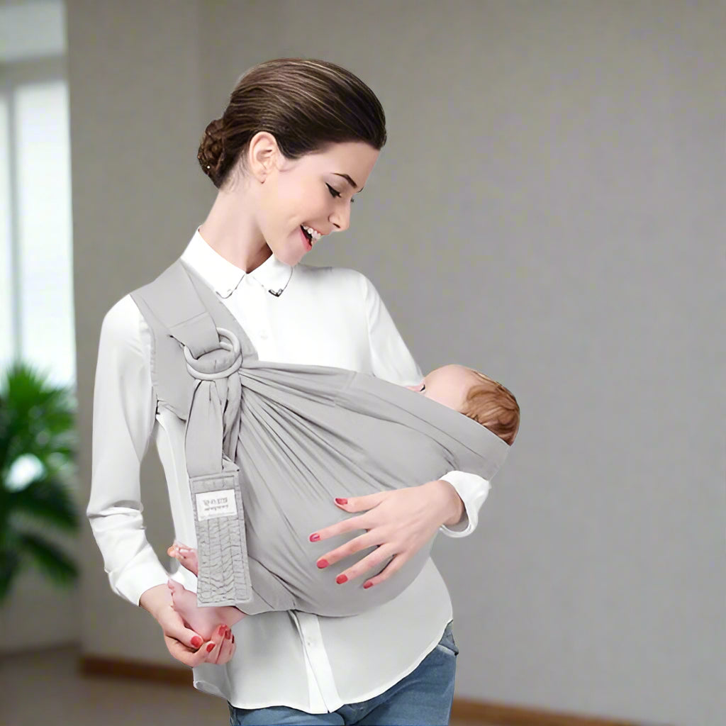 Une femme en chemisier blanc tient un bébé dans le portage Echarpe | SwaddleSling™, souriant au bébé avec un mur neutre et une plante verte en arrière-plan.