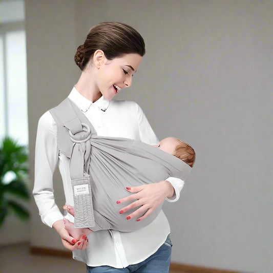 Une femme en chemisier blanc sourit à son bébé, blotti dans un porte-bébé gris clair Echarpe Portage | SwaddleSling™. Elle se tient à l'intérieur, devant un arrière-plan uni et une plante verte visible.
