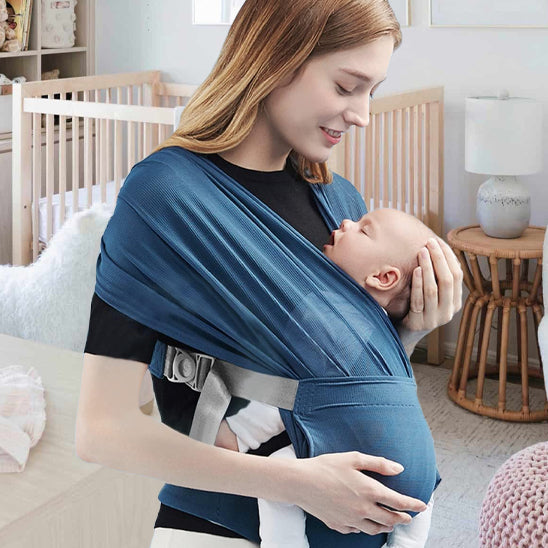 Une femme porte l'Écharpe porte-bébé | WrapTight™ bleue pour serrer son bébé endormi contre elle, créant ainsi un lien unique. Elles se trouvent dans une chambre d'enfant à la lumière tamisée, avec un berceau, une table en osier et une chaise moelleuse en arrière-plan.