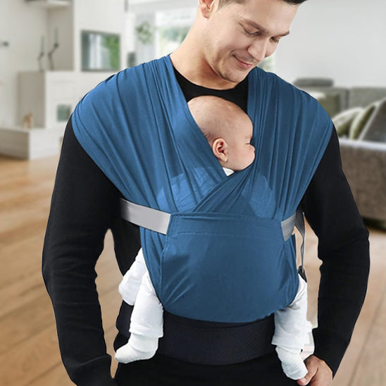 Un homme, debout à l'intérieur, vêtu d'une chemise noire, tient un bébé dans ses bras grâce à l'écharpe porte-bébé bleue WrapTight™, pour un portage mains libres. Le bébé est tourné vers l'intérieur, avec un salon moderne en arrière-plan.