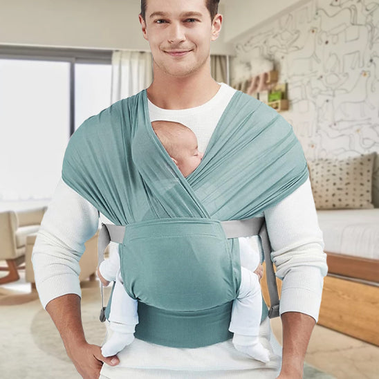 Un homme porte l'Écharpe porte-bébé bleu clair | WrapTight™ et tient son bébé endormi contre sa poitrine. Grâce à sa conception « mains libres », il profite du confort d'un lit, de chaises et d'une fenêtre à rideaux en arrière-plan.