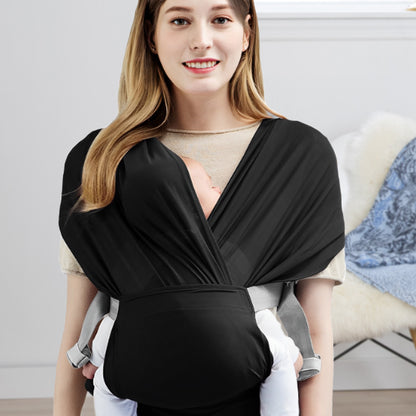 Une femme aux longs cheveux blonds sourit, vêtue d'un haut beige et portant son bébé les mains libres dans une écharpe porte-bébé noire | WrapTight™. La pièce présente une décoration neutre avec une chaise blanche et une couverture bleue en arrière-plan.